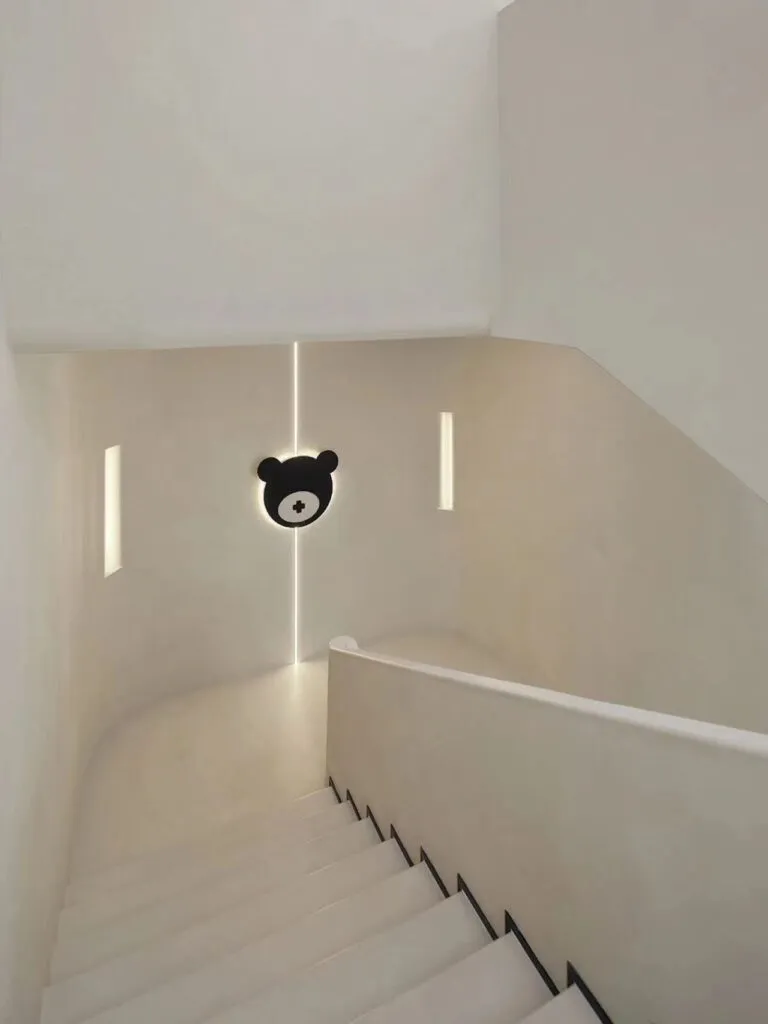 Minimalist cream-toned staircase landing featuring smooth seamless walls, subtle vertical strip lighting, and a black bear emblem on the wall
