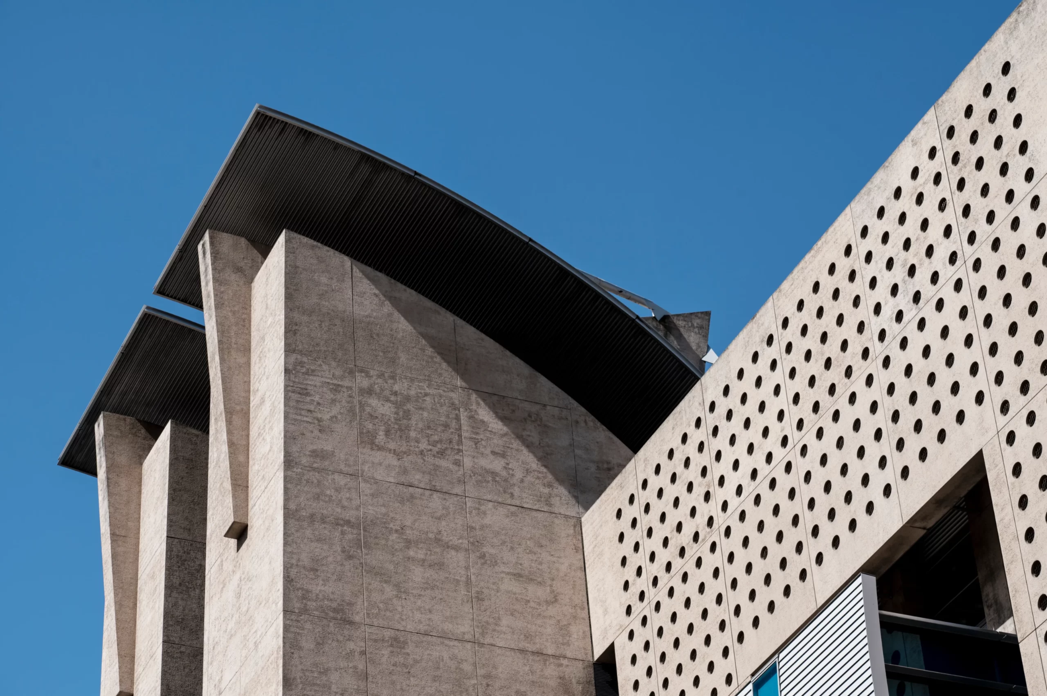 Modern architectural concrete facade with curved roof overhangs and a perforated wall against a clear blue sky
